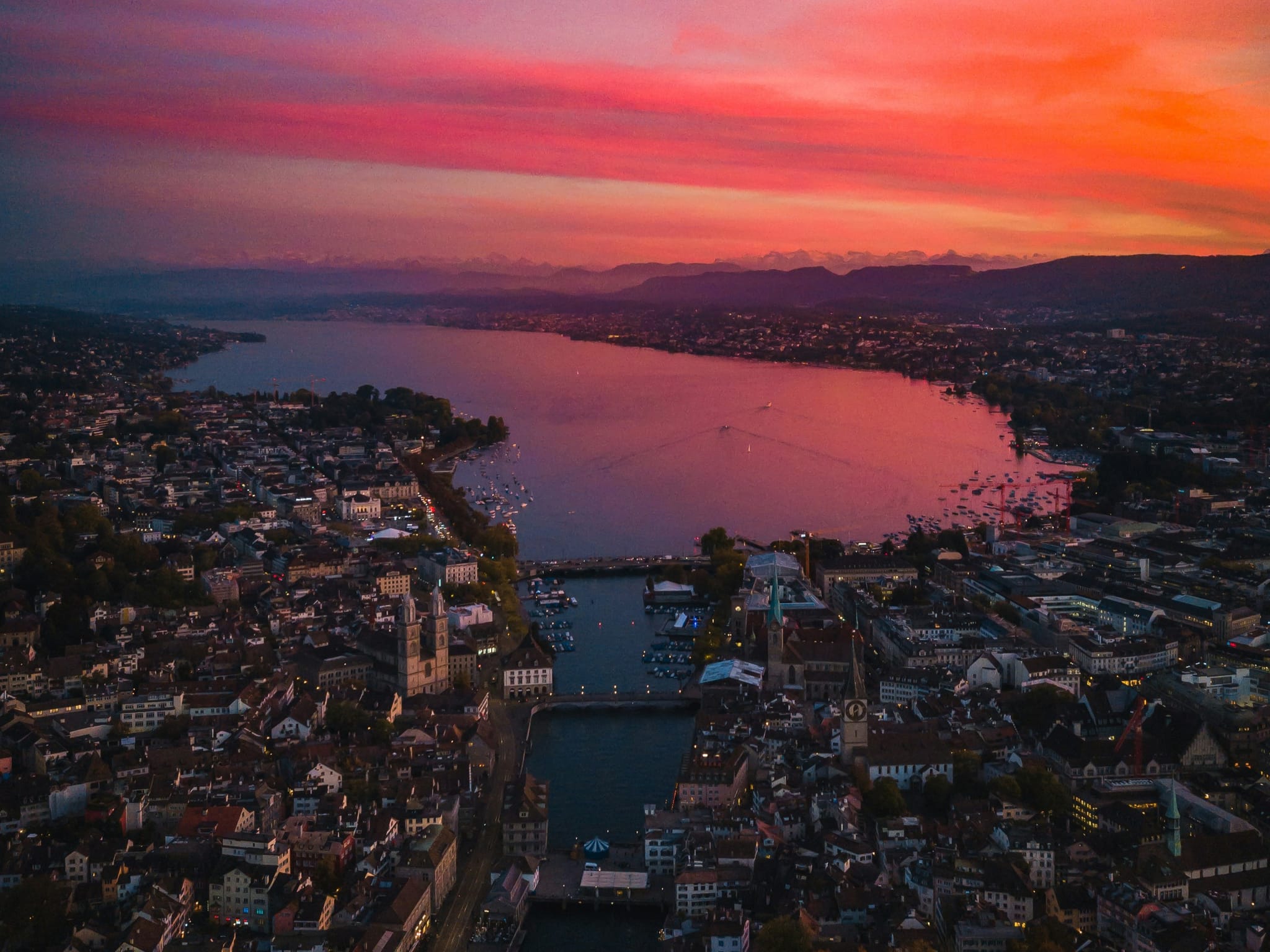 Luftaufnahme der Abenddämmerung von Zürich und dem Zürichsee mit dem Fluss Limmat, Bergen in der Ferne und verbleibenden roten Sonnenuntergangslichtern, die eine dramatische Alpenabendlandschaft schaffen.